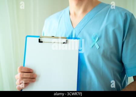 Male healthcare professional in uniform with blue ribbon symbol of prostate cancer awareness holding clipboard with empty paper for notes and reminders in November. Stock Photo