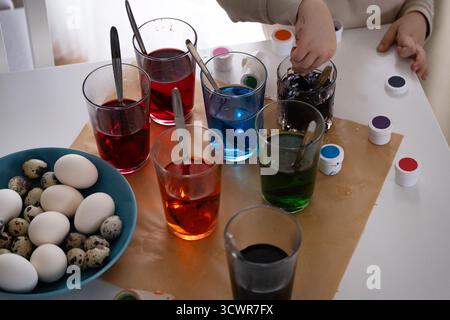 Close-up of freshly dyed Easter eggs resting on a soft cloth near glasses with red and yellow paint. The scene captures the cozy atmosphere of home Ea Stock Photo