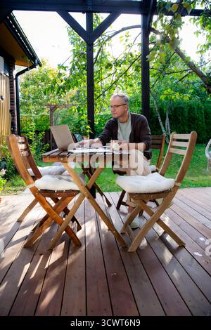 Middle aged man working remotely on laptop at cozy sunny patio, digital nomad lifestyle, outdoor home office, freelance and productivity concept Stock Photo