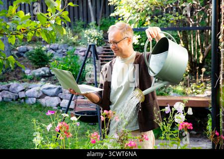 Middle-aged man watering flowers and working remotely on laptop during video call on sunny patio, modern multitasking lifestyle concept. Stock Photo