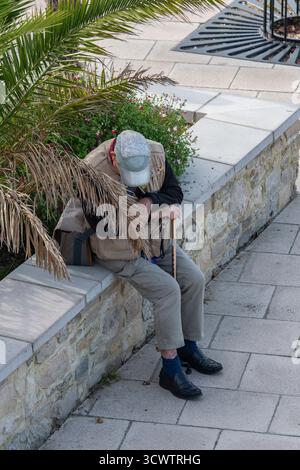 old or retired man sitting on a wall taking a rest holding a walking stick. Stock Photo