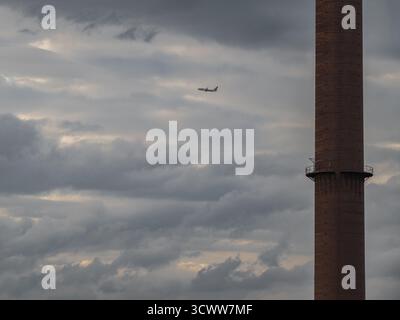Airplane Flying Past Industrial Chimney under Dramatic Cloudy Sky Symbolizing Travel and Industry Stock Photo