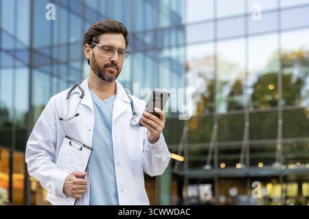 Male doctor wearing scrubs and a white coat, holding a clipboard and smartphone outside a modern clinic, using mobile technology for healthcare communication Stock Photo