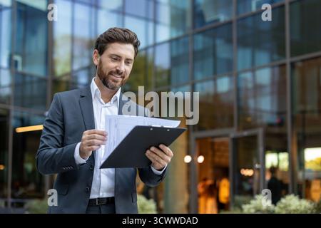 Confident businessman reviewing important papers on a clipboard with a content smile, standing in front of a modern glass office building, symbolizing success and corporate life Stock Photo