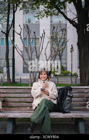 Woman sitting on a park bench smiling while using a smartphone with a backpack. Concept of outdoor relaxation and digital connectivity Stock Photo