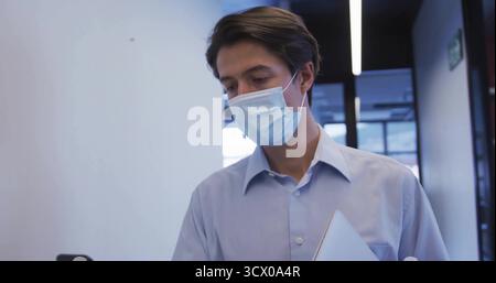Walking man wearing shirt and mask, checking smartphone, holding laptop in office corridor Stock Photo