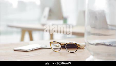 Resting tortoiseshell reading glasses by smartphone on office desk with blurred monitor pitcher Stock Photo