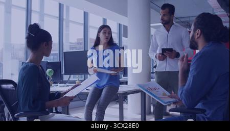 Collaborating office colleagues reviewing printed reports and charts at office desks, with tablet Stock Photo