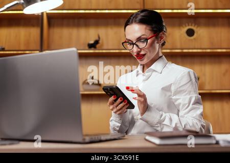 Professional woman checks messages on phone while working on a laptop, ideal for mobile app, communication, login, verification and customer workflow Stock Photo