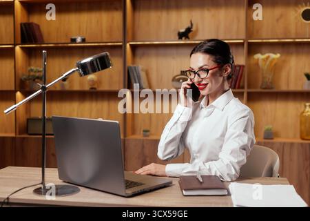 Smiling professional talks on a smartphone while working on a laptop, illustrating communication, sales, customer support and remote work in a modern Stock Photo