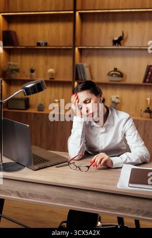 Tired office worker holds head with glasses beside laptop, illustrating migraine, stress and screen fatigue in the workplace. Stock Photo