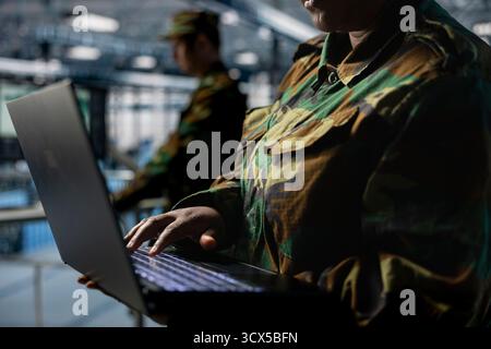 Close up of server hub employee in soldier uniform typing on laptop, running diagnostics on government systems. Army information technology specialist in data center writing code on notebook keyboard Stock Photo