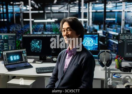 Smiling server hub senior programmer using devices to monitor neural network with AI LLM. Asian man at work in data center using artificial intelligence to oversee servers powering machine learning Stock Photo