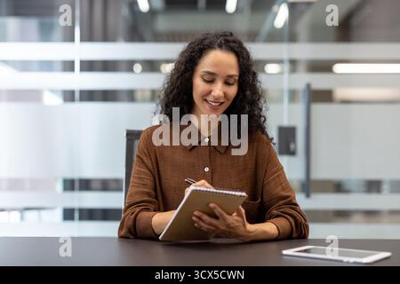 Woman with curly hair sitting at a modern office desk, actively taking notes in a spiral notebook, showing a pleasant and engaged expression during a business meeting or while working Stock Photo