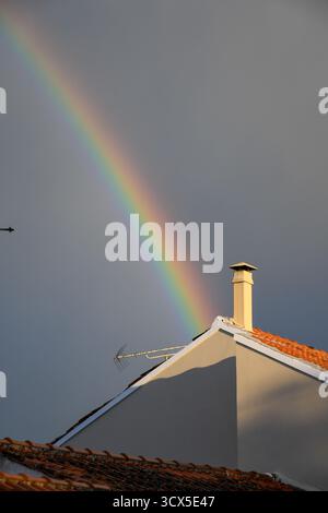 Vibrant rainbow arc ending behind a sunlit rooftop with a chimney against a dark, stormy sky. A beautiful natural weather phenomenon after rain in a E Stock Photo
