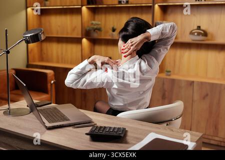 Back view of a woman stretching her neck at a laptop desk, illustrating office ergonomics, microbreaks, posture care and relief from stiffness. Stock Photo