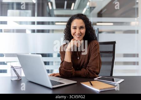 Confident young businesswoman smiling and looking at the camera, posing at a modern office desk with a laptop, reflecting professionalism and success in a corporate setting Stock Photo
