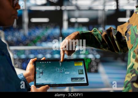 Military technician reviewing tactical dashboard on tablet screen in server hub, standing on industrial platform. Army expert using strategic communications and infrastructure software systems. Stock Photo