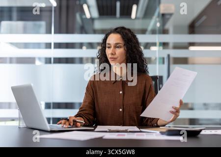 Young professional businesswoman managing corporate tasks, inputting data into a laptop while reviewing important financial documents and contracts at an office desk Stock Photo