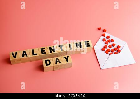 Flat lay composition with wooden blocks spelling “Valentine Day” and a white envelope filled with small red hearts on a pink background. Stock Photo