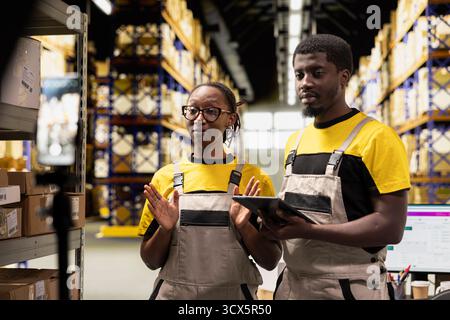 Coworkers collaborating on a live stream in storage room warehouse, showcasing parcel handling and order processing. African American workers present goods for a commercial campaign. Stock Photo