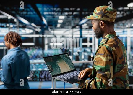 African american military staff reviewing tactical imagery on laptop in server hub control center, standing on industrial platform. Army officer using strategic geospatial data for security. Stock Photo