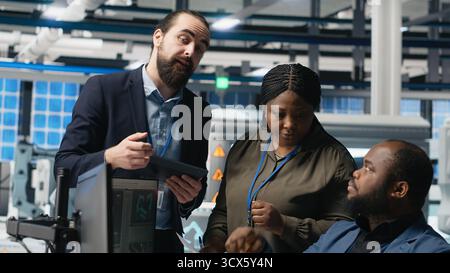 Multiethnic master engineers doing teamwork on high tech production line for solar panels manufacturing activity, combining technical insight and collaboration for green energy. Camera B. Stock Photo