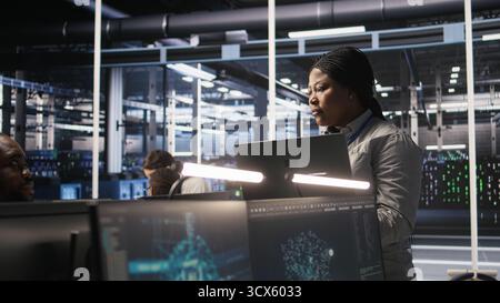 Data center manager supervising engineer developing software to analyze neural networks at computer workstation. Team leader and employee observing system performance in AI server room, camera A Stock Photo