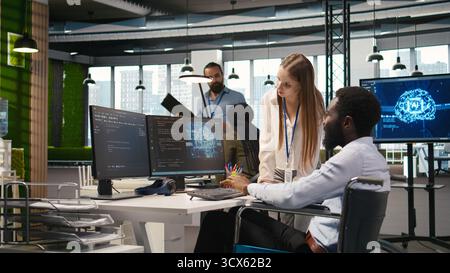 Junior dev and coworker in wheelchair developing software applications, optimizing code. Female technician and colleague with paraplegia in office using AI to solve bottlenecks, camera A Stock Photo