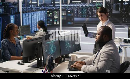 Happy data center manager providing positive feedback to engineer developing neural networks software on computer. Cheerful supervisor pleased by employee work implementing AI in server room, camera A Stock Photo