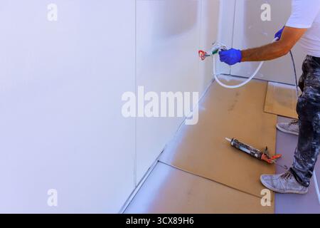 Skilled worker uses spray tool to apply sealant on wall, ensuring seamless finish in room workspace. Stock Photo