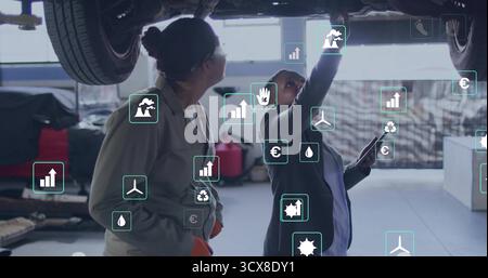 Using tablet, two female mechanics in coveralls inspecting car underbody in workshop, overlay icons Stock Photo