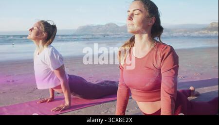 Practicing two women in sportswear holding cobra poses on sandy beach, with colorful yoga mats Stock Photo