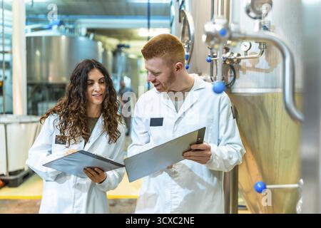 Two brewery workers in lab coats inspecting machinery and documenting processes, ensuring product quality and efficient operation in a modern industri Stock Photo