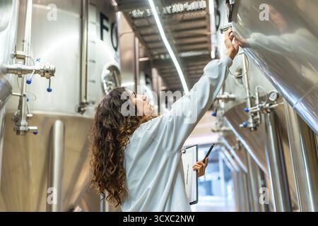Young woman in a lab coat checking control panel on a stainless steel fermentation tank, performing quality control and inspection during the beer pro Stock Photo