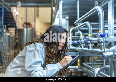Woman wearing a lab coat meticulously checking industrial pipes and valves in a modern beverage factory, performing a detailed quality control inspect Stock Photo
