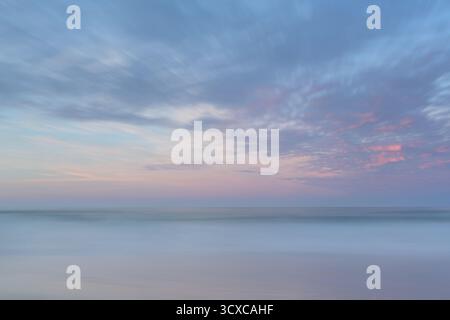 Pastel hues of pink and blue fill the clouds, reflecting the gentle light of sunset over a misty beach. The horizon is a soft blend of sky and ocean. Stock Photo
