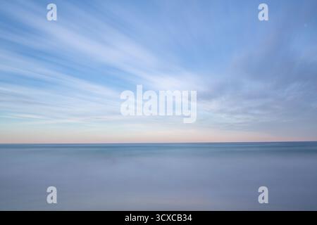 Wispy clouds stretch across a serene blue sky above a calm, misty seascape. The horizon separates the soft blue tones of the sky from the gentle hues. Stock Photo
