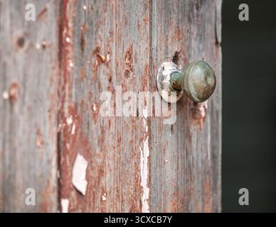 Weathered wooden door with peeling paint showing a mix of aged red and gray hues. A round, tarnished brass doorknob is set on a chipped white plate. T Stock Photo