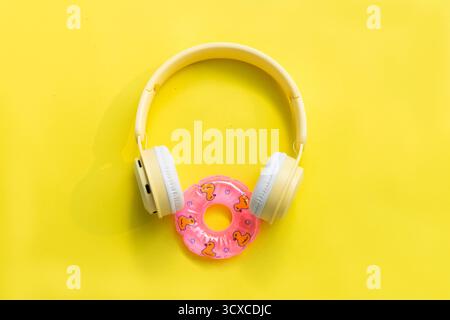 Stylish yellow headphones placed on a pink inflatable swim ring, isolated on a yellow background. Trendy flat lay concept for summer music, vacation, Stock Photo
