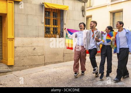 Happy queer friends celebrating lgbtq pride on a sunny historic cobblestone street in madrid, holding a rainbow flag, fan and megaphone, smiling, unit Stock Photo