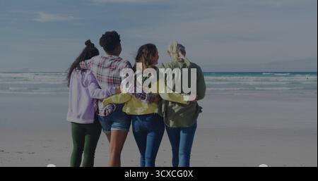 Hugging friends wearing hoodies, plaid shirts, jackets and jeans on sandy beach, with sea view Stock Photo