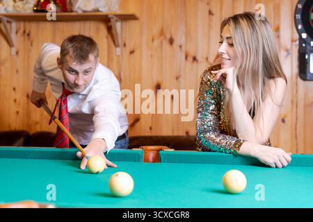 Man is playing pool with a woman. The man is wearing a red tie. The woman is sitting on the side of the pool table Stock Photo