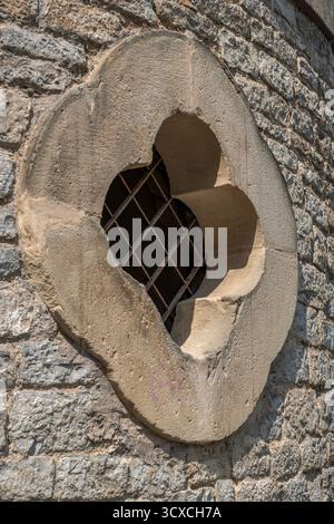 Close-up of an ancient stone window detail with a stylized trifoliate or quadrifoil pattern and a protective metal grille on a weathered medieval wall Stock Photo