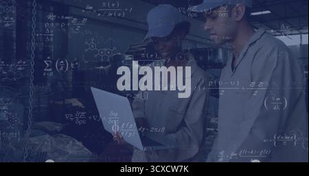 Analyzing data, technicians in coveralls monitoring laptop on plant floor, with drums, shelving Stock Photo