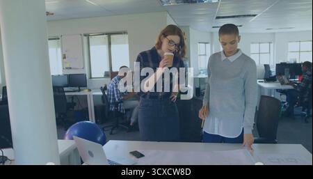 Reviewing printed blueprint two female colleagues at white table in office, with coffee cup pencil Stock Photo