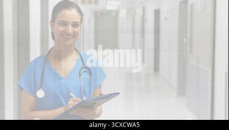Writing Indian nurse wearing blue scrubs with stethoscope in hospital hall with clipboard and pen Stock Photo
