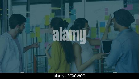 Collaborating coworkers holding laptop pointing at colorful sticky notes on glass wall at office Stock Photo