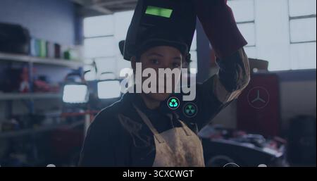 Female welder raising welding helmet overhead in workshop, with leather apron and protective gloves Stock Photo