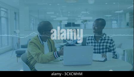 Two colleagues discussing project while gesturing toward laptop in open-plan office, with paperwork Stock Photo